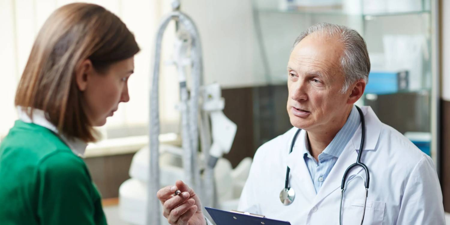 Senior doctor discussing workplace wellness and chiropractic care with a patient, holding a pen and notepad in a clinic setting.
